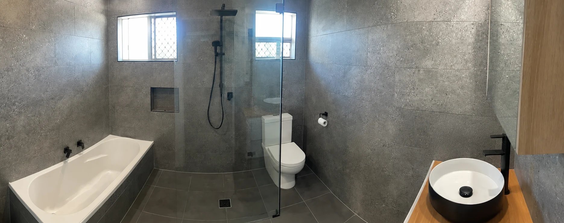Modern bathroom interior with white soaking tub on left, frameless glass shower enclosure with toilet in center, and round vessel sink on wooden counter on right