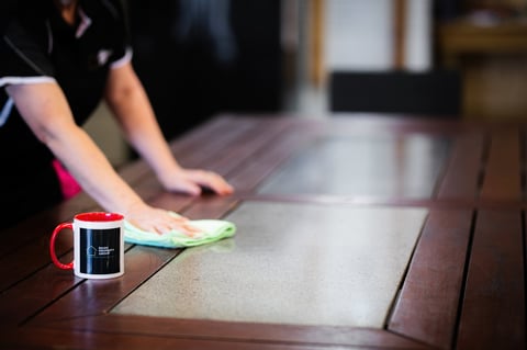 Person in black shirt wiping wooden table with cloth next to red mug and black mug