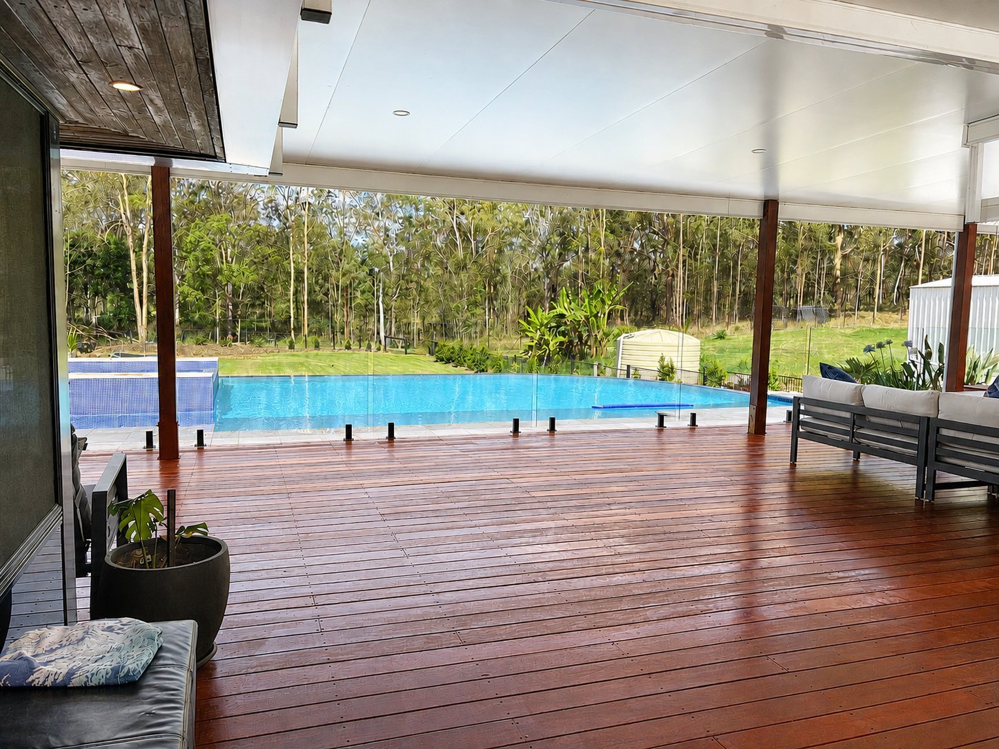 Modern covered deck with red timber flooring overlooking a bright blue swimming pool and wooded landscape