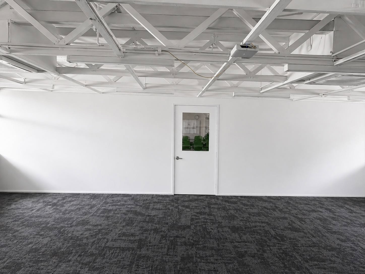 Empty industrial warehouse interior with exposed ceiling beams, white walls, gray carpet flooring, and a single white door with small window