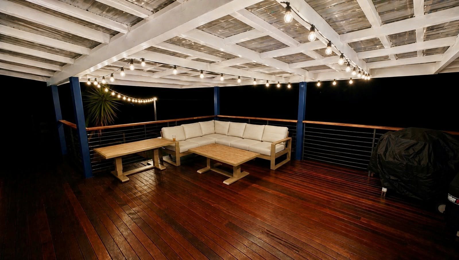 Rooftop seating area at night with white cushioned L-shaped bench, wooden deck, string lights, and dark railings