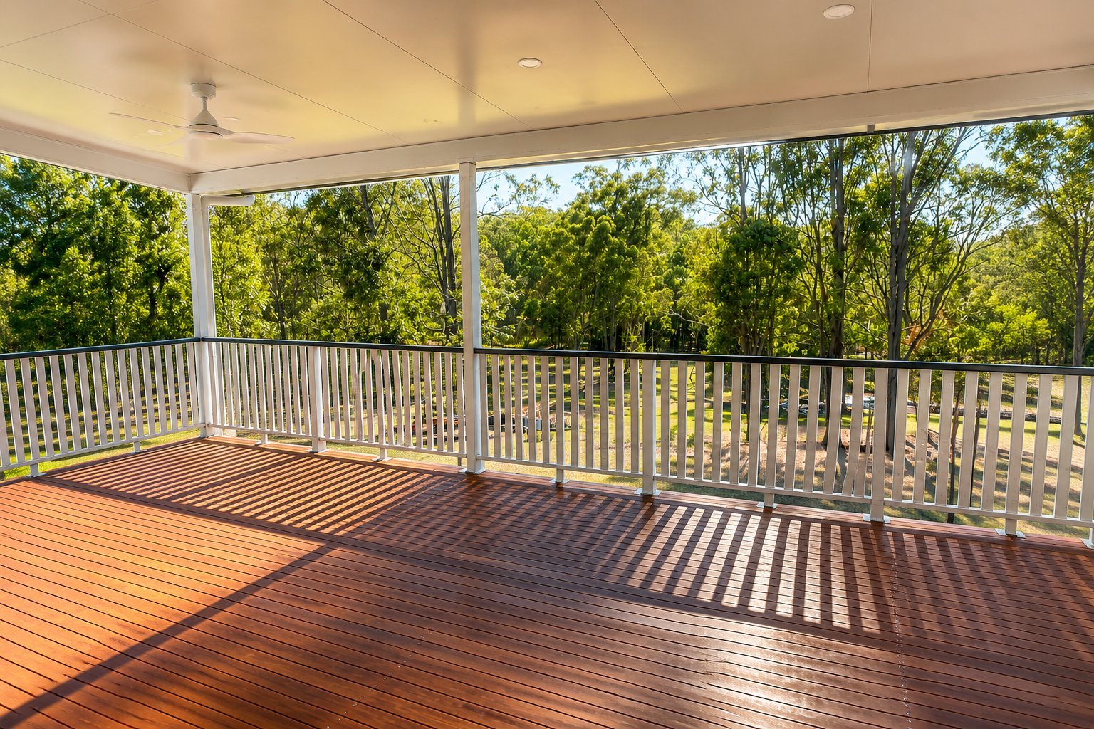Empty covered deck with brown composite flooring, white railings, and views of green trees and sky through large windows
